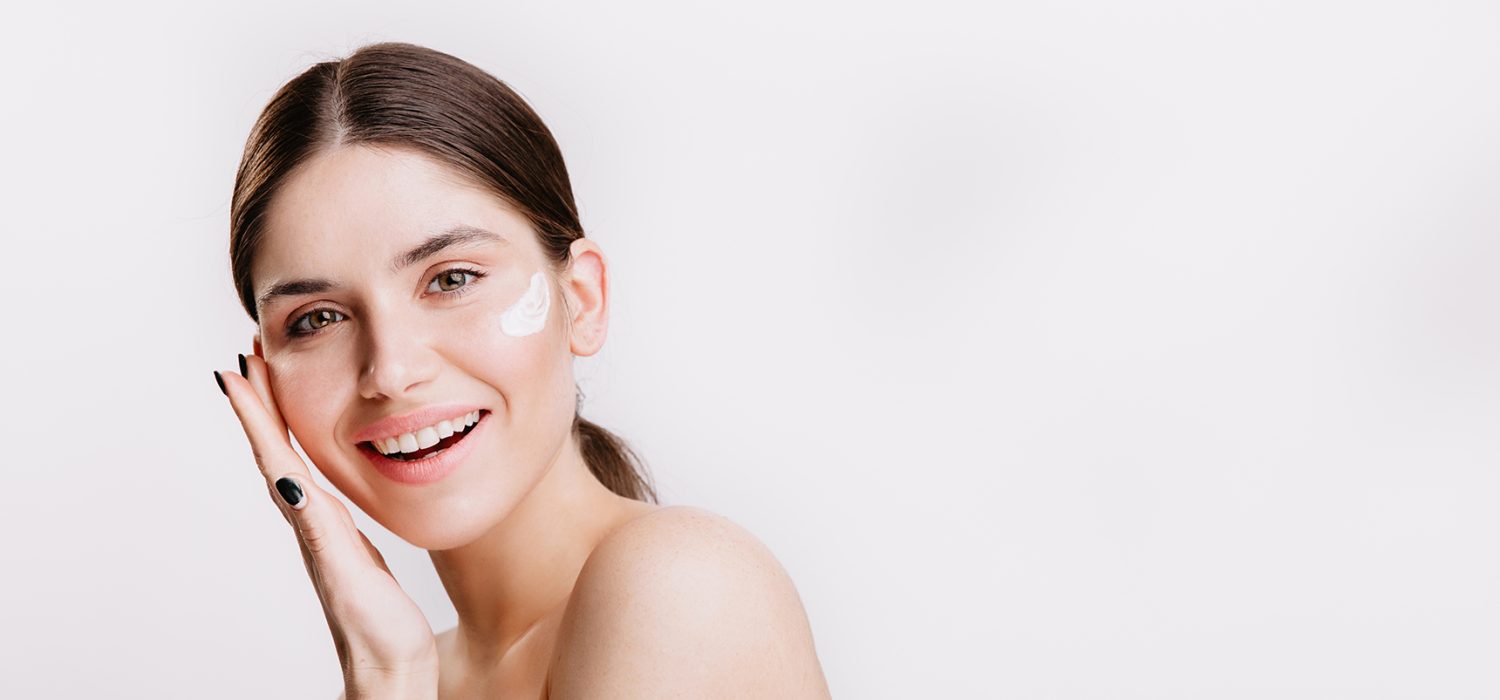 Girl touches moisturized skin and smiles. Portrait of model with cream on face on isolated background